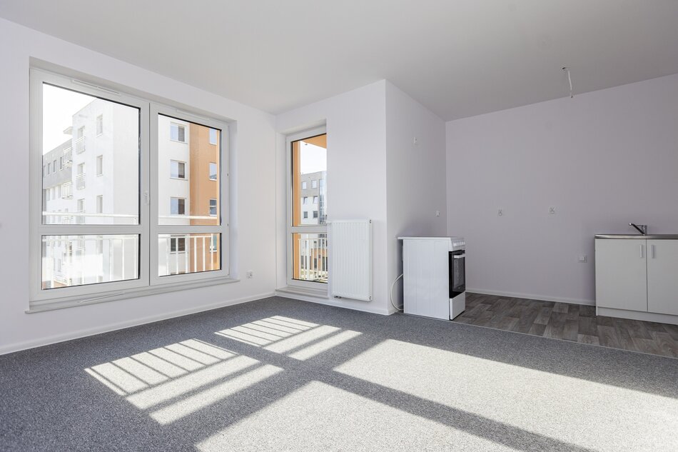 na zdjęciu fragment nowego mieszkania, po lewej widać dwa okna, białe ściany, szarą wykładzinę na podłodze, po prawej biała kuchenka i zlewozmywak