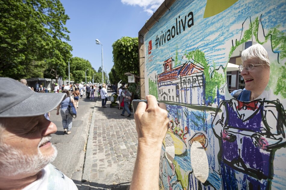 Na zdjęciu widać starszego mężczyznę robiącego zdjęcie starszej kobiecie, która wygląda przez wycięcie w dużej, kolorowej tablicy dekoracyjnej z napisem #vivaoliva. W tle znajduje się tłum ludzi spacerujących po wybrukowanej ulicy otoczonej drzewami