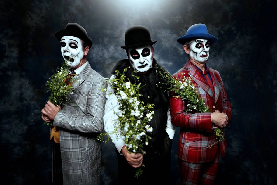 Na zdjęciu znajduje się trzech mężczyzn z twarzami pomalowanymi na biało-czarno w stylu mimów, ubranych w eleganckie garnitury i kapelusze, każdy z nich trzymający bukiet kwiatów. Tło jest ciemne i nastrojowe, co dodaje zdjęciu dramatycznej atmosfery.