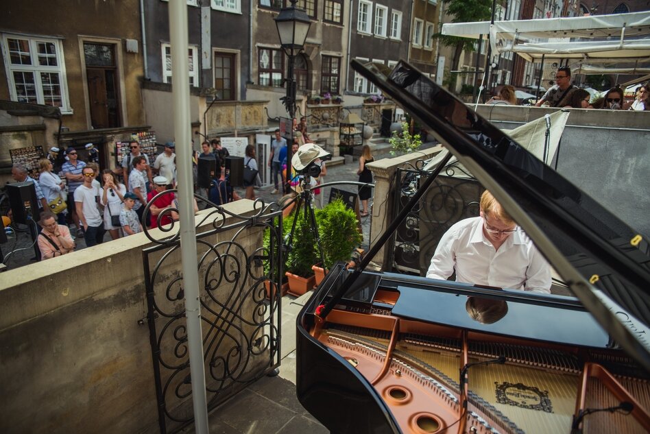 Zdjęcie przedstawia pianistę grającego na fortepianie na zewnątrz, w otoczeniu zabytkowej zabudowy miejskiej. W tle widać przechodniów oraz publiczność, która przysłuchuje się koncertowi