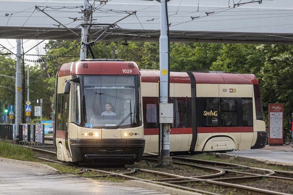 Na zdjęciu znajduje się nowoczesny tramwaj typu Swing jadący po torach tramwajowych, z numerem bocznym 1029. Tramwaj ma czerwono-kremowe barwy i znajduje się na otwartej przestrzeni, prawdopodobnie w pobliżu przystanku, z zielonymi drzewami w tle i wiaduktem nad torami.