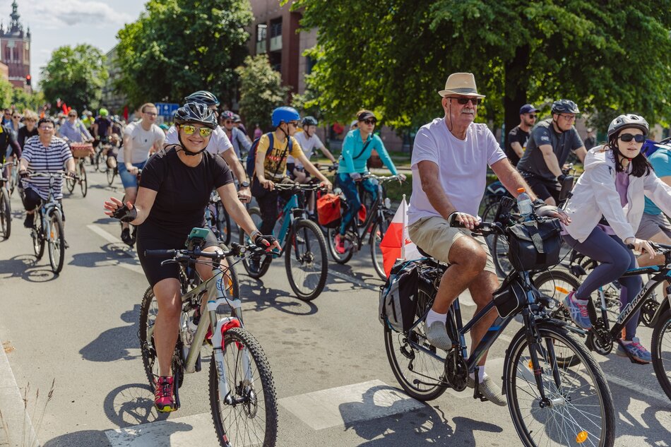 Na zdjęciu widzimy grupę rowerzystów uczestniczących w przejeździe ulicznym, jadących w pogodny dzień. Wśród uczestników znajdują się osoby w różnym wieku, w tym kobieta w ciemnych okularach oraz mężczyzna w jasnym kapeluszu, co dodaje zdjęciu różnorodności i przyjaznej atmosfery.