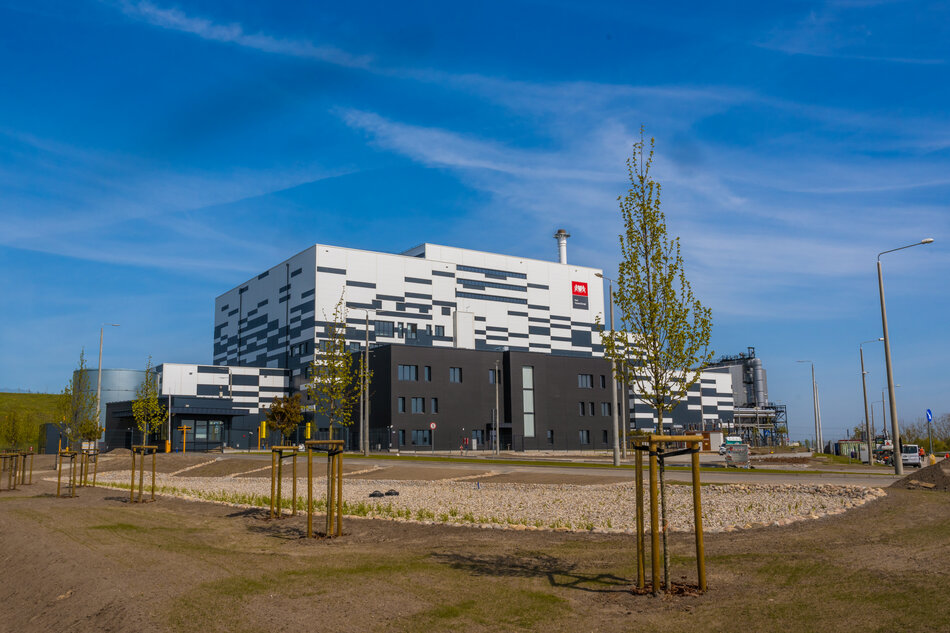 Na zdjęciu znajduje się nowoczesny budynek przemysłowy oznaczony jako Port Czystej Energii. Budynek ma biało-czarne, geometryczne elewacje i jest otoczony nowo nasadzonymi drzewami oraz uporządkowanym terenem