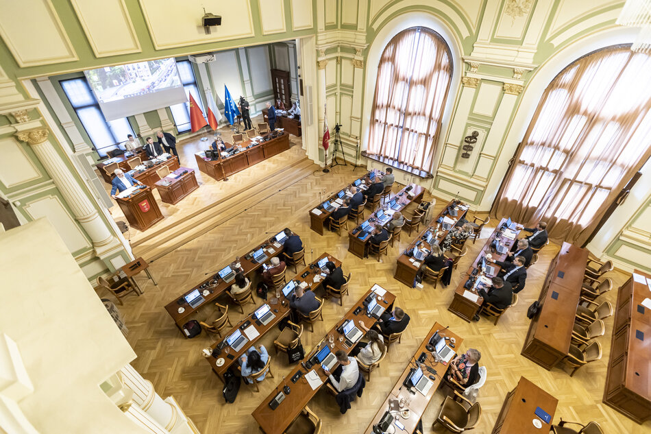 Na zdjęciu przedstawiona jest sesja Rady Miasta, odbywająca się w eleganckiej sali z wysokimi oknami i dekoracyjnymi ścianami. Radni siedzą przy drewnianych stołach wyposażonych w laptopy, a na podwyższeniu znajduje się prezydium oraz stanowisko z flagami Polski i Unii Europejskiej.