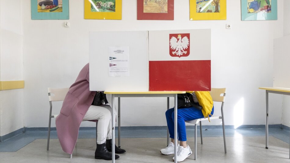 Zdjęcie przedstawia dwie osoby siedzące przy stole z przegrodą do głosowania w pomieszczeniu wyborczym. Przegroda ma biało-czerwoną flagę z orłem, co sugeruje, że miejsce to znajduje się w Polsce. Obie osoby siedzą na plastikowych krzesłach, a ich górne partie ciała są zasłonięte przez przegrodę. Osoba po lewej stronie nosi różowy płaszcz i białe spodnie, a osoba po prawej stronie ma na sobie żółtą kurtkę i niebieskie spodnie. W tle widać kilka kolorowych obrazków powieszonych na ścianie.