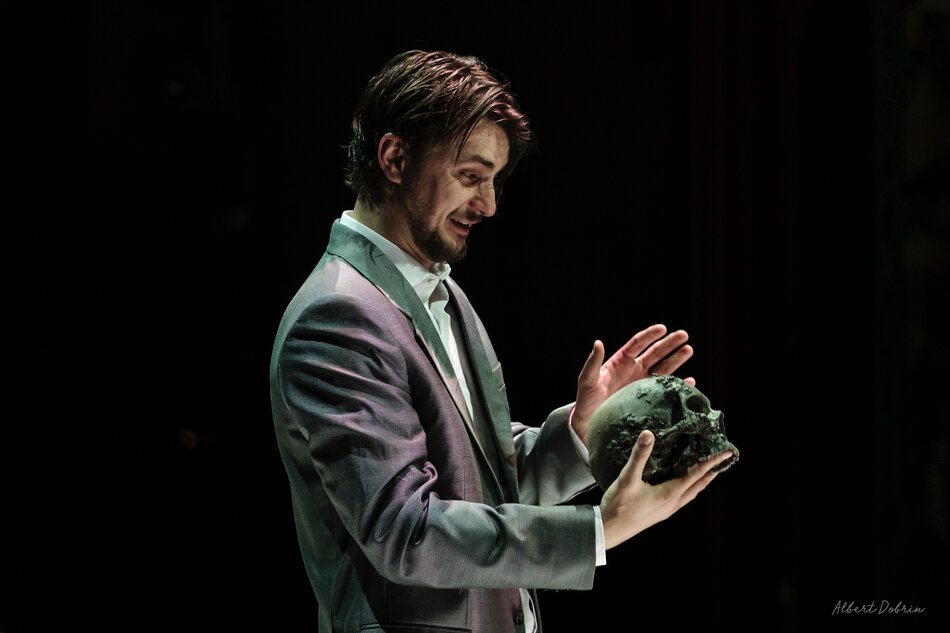 Zdjęcie przedstawia mężczyznę w garniturze, który trzyma czaszkę w dłoniach, co jest nawiązaniem do słynnej sceny z Hamleta Williama Szekspira
