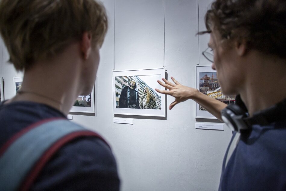 Na zdjęciu widać dwóch mężczyzn oglądających zdjęcia wiszące na ścianie w galerii. Jeden z nich wskazuje na jedną z fotografii, a drugi uważnie się jej przygląda