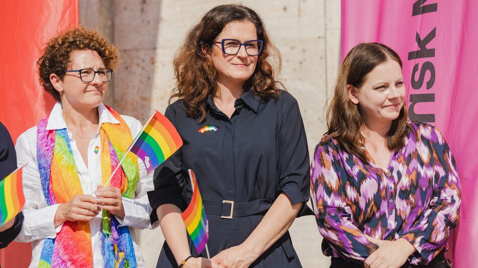 Zdjęcie przedstawia trzy kobiety stojące obok siebie na zewnątrz, uczestniczące w wydarzeniu związanym z ruchem LGBTQ+. Wszystkie kobiety mają przypięte tęczowe symbole lub trzymają tęczowe flagi.Kobieta po lewej stronie ma krótkie kręcone włosy, nosi okulary i białą koszulę z kolorowym, tęczowym szalem. Kobieta pośrodku ma ciemne, falowane włosy i ciemne okulary. Ubrana jest w ciemną sukienkę z tęczową przypinką. Kobieta po prawej ma proste, ciemne włosy i ubrana jest w kolorową, wzorzystą bluzkę. W tle widać jasne, kamienne ściany oraz różowy baner z czarnym napisem.