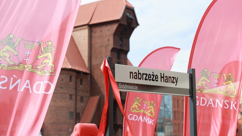 Zdjęcie przedstawia uliczny znak nabrzeże Hanzy w Gdańsku na tle charakterystycznej architektury ceglanej. Obok znaku widoczne są czerwone flagi z herbem Gdańska, na których widnieją złote lwy trzymające tarczę z krzyżem.