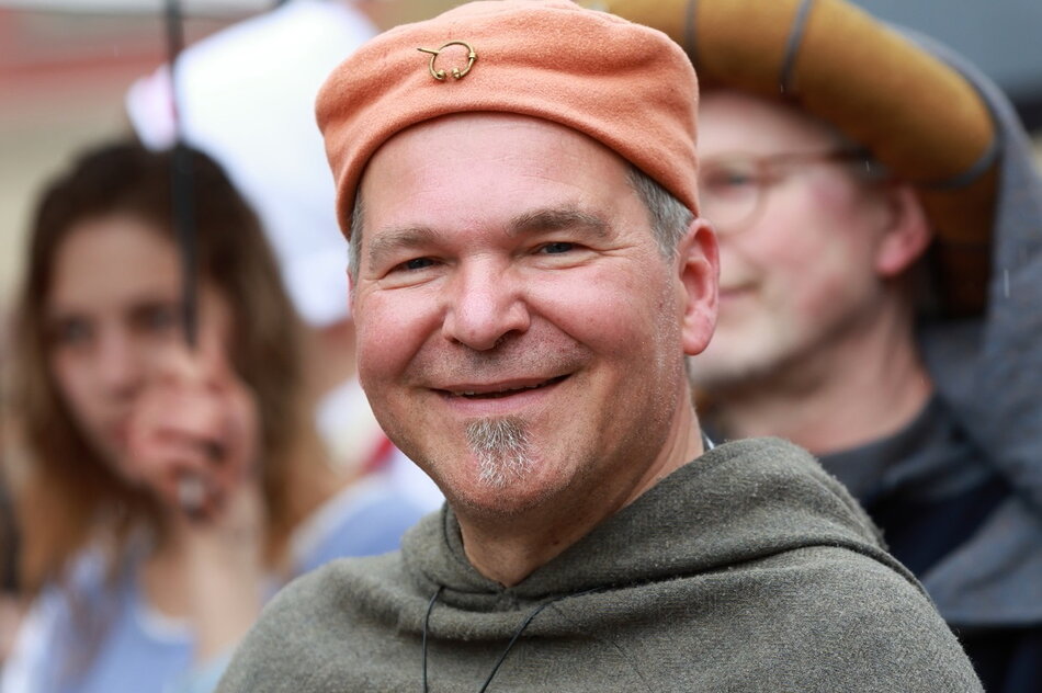 Na zdjęciu widać uśmiechniętego mężczyznę w średniowiecznym stroju, noszącego brązowy beret i medalion na szyi. W tle widać innych uczestników wydarzenia, którzy również są ubrani w historyczne kostiumy, co sugeruje, że biorą udział w rekonstrukcji historycznej lub festiwalu.