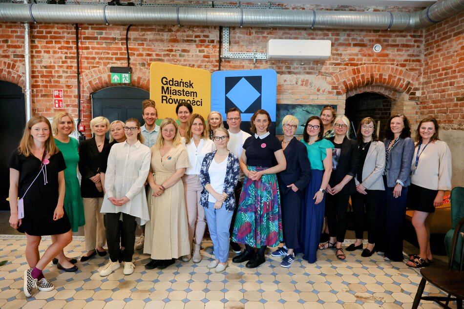 Na zdjęciu widzimy grupę ludzi pozujących do wspólnego zdjęcia w jasnym pomieszczeniu z ceglaną ścianą w tle. W tle znajdują się banery z napisem Gdańsk Miastem Równości, co sugeruje, że wydarzenie jest związane z promocją równości i integracji społecznej.