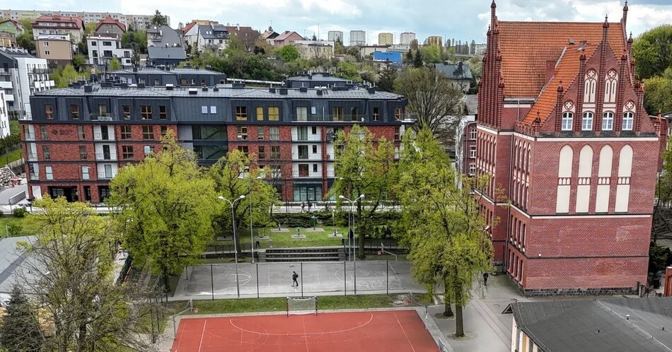 Na zdjęciu widoczny jest kompleks budynków w Siedlcach, z nowoczesnym, wielopiętrowym budynkiem mieszkalnym po lewej stronie i zabytkowym budynkiem z czerwonej cegły po prawej stronie. Przed budynkami znajduje się boisko sportowe otoczone drzewami, a w tle widać mieszkalne zabudowania na wzgórzu.