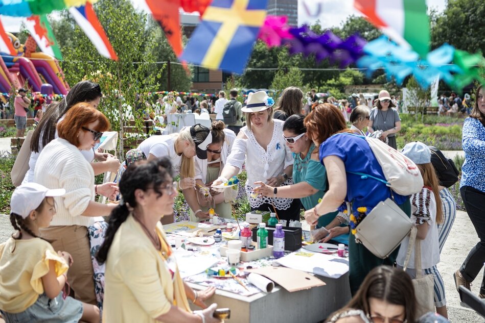 Na zdjęciu widać grupę osób uczestniczących w warsztatach plastycznych na świeżym powietrzu, przy stoliku pełnym farb, pędzli i innych materiałów artystycznych. W tle znajdują się kolorowe dekoracje, girlandy z flagami oraz dmuchane atrakcje, a wokół krąży wiele osób, tworząc atmosferę pikniku rodzinnego.