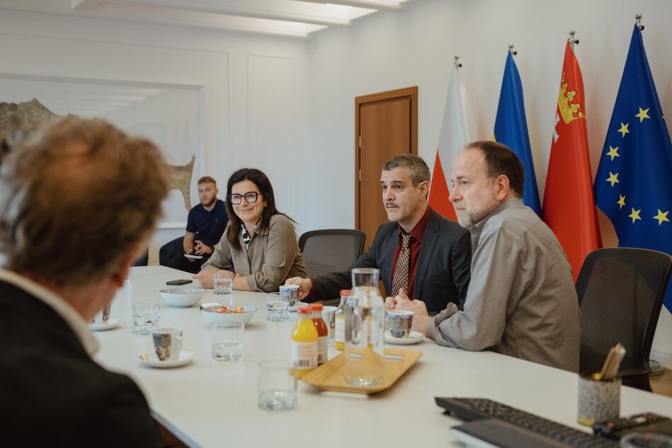Zdjęcie przedstawia grupę ludzi siedzących przy stole konferencyjnym, na którym znajdują się napoje i przekąski. W tle widać flagi Polski, Unii Europejskiej oraz innych regionów, co sugeruje, że spotkanie ma charakter oficjalny