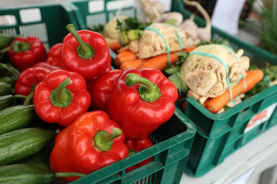 na zdjęciu czerwona papryka w zielonej skrzyni, widać kilka tych warzyw, obok leży marchewka połączona z selerem i innymi białymi warzywami, także w zielonej skrzyni