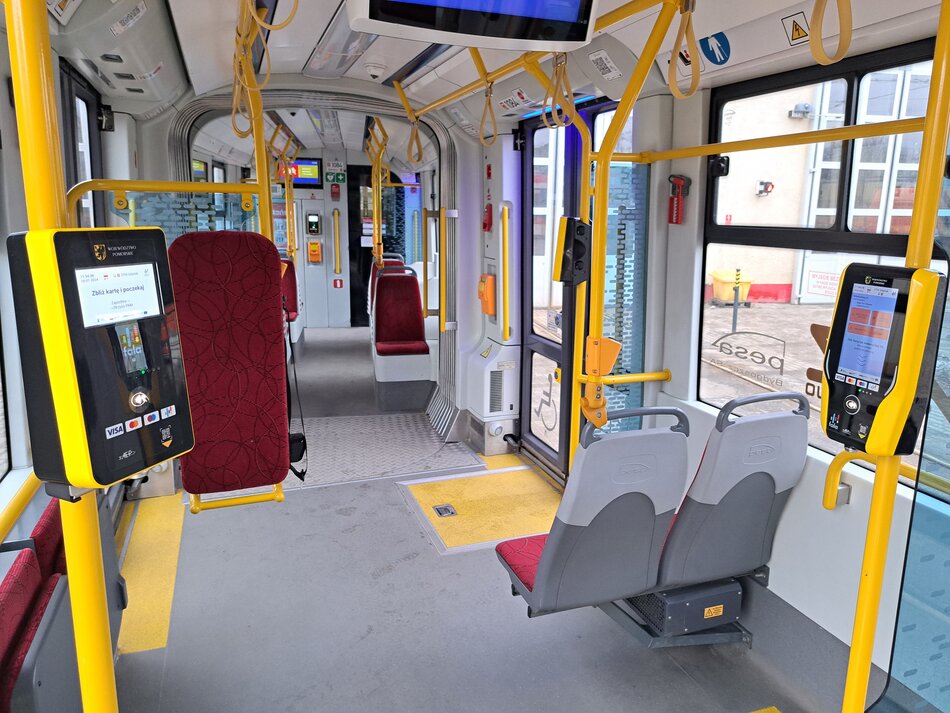 Wnętrze autobusu miejskiego, są krzesełka dla pasażerów, po lewej widać urządzenie na słupku do kupowania biletów