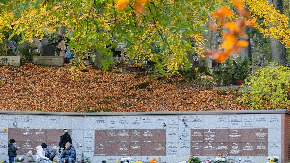 Na zdjęciu widać kolumbarium, przed którym kilka osób składa kwiaty i zapala znicze. W tle widoczny jest cmentarz z nagrobkami, otoczony jesiennymi drzewami i opadłymi liśćmi.