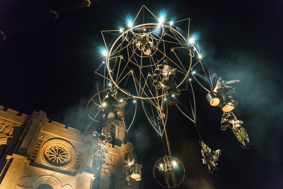 Zdjęcie przedstawia spektakl nocny na świeżym powietrzu, gdzie artyści z Compagnie Transe Express wykonują akrobacje i grają na instrumentach, zawieszeni na skomplikowanej metalowej konstrukcji przypominającej ogromną karuzelę. W tle widać oświetloną fasadę gotyckiego budynku, co dodaje scenie dramatyzmu i magii