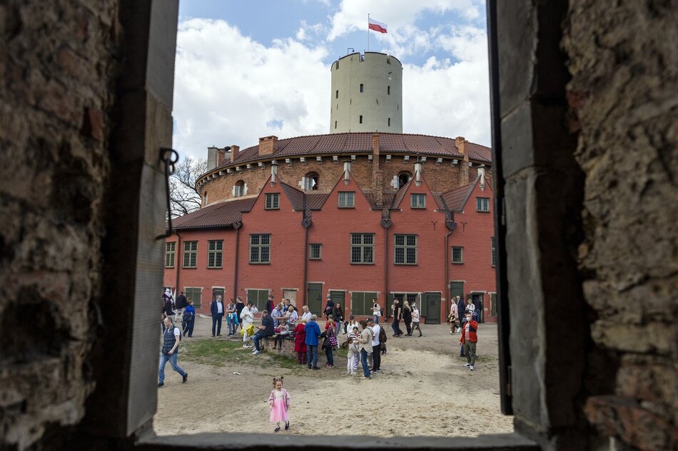 Zdjęcie przedstawia widok na Twierdzę Wisłoujście w Gdańsku, oglądaną przez okno ruin. Widać tłum ludzi zgromadzonych na dziedzińcu twierdzy, a w tle wznosi się charakterystyczna wieża z polską flagą na szczycie.