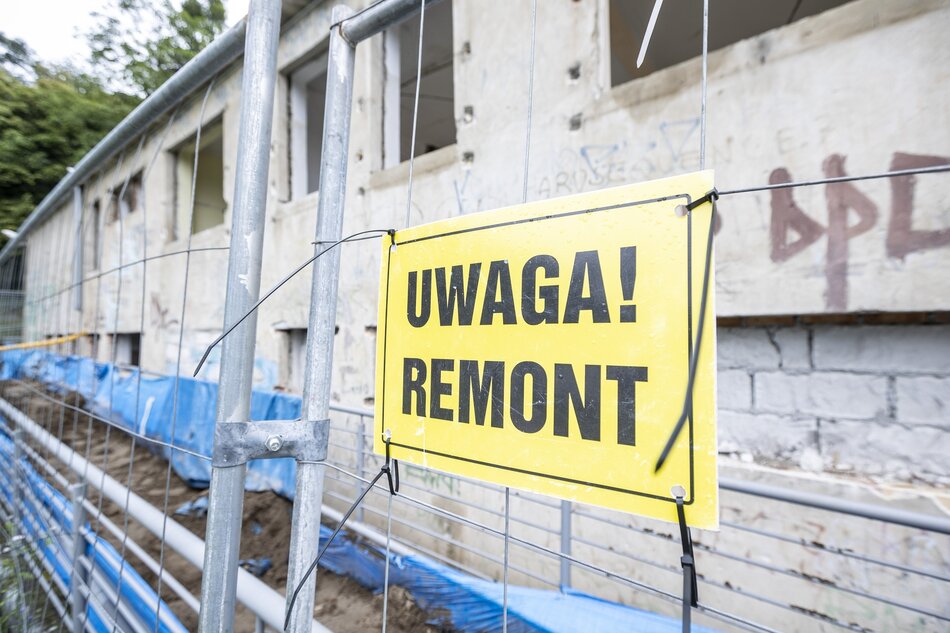 Zdjęcie przedstawia budynek w trakcie remontu, otoczony metalowym ogrodzeniem. Na ogrodzeniu widnieje żółta tablica z napisem UWAGA! REMONT, ostrzegająca przechodniów o trwających pracach.