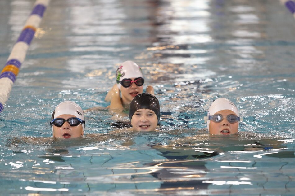 Na zdjęciu widać cztery młode osoby w basenie, wszystkie mają na głowach czepki pływackie i okulary do pływania. Osoby są w trakcie pływania, a każda z nich jest widoczna na powierzchni wody z wyraźnymi uśmiechami na twarzach.