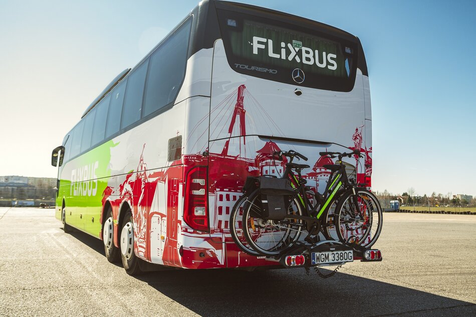 Na zdjęciu znajduje się autokar FlixBus marki Mercedes-Benz Tourismo, zaparkowany na otwartej przestrzeni. Na tylnym bagażniku zamontowane są dwa rowery, a tylna część autobusu jest ozdobiona grafiką przedstawiającą miasto w czerwonych barwach.