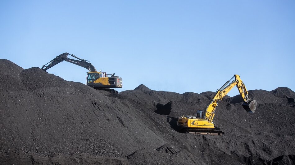 Zdjęcie przedstawia dwie koparki pracujące na hałdach czarnego materiału, prawdopodobnie węgla. Jedna koparka jest żółta, druga natomiast czarna. Obie maszyny są w trakcie przenoszenia materiału, co można zauważyć po ułożeniu ich łyżek. W tle widoczny jest jasnoniebieski niebo, a teren wokół koparek to rozległe, nierówne hałdy węgla