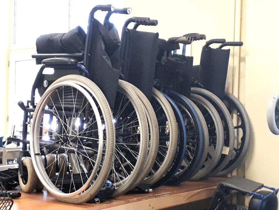 Na zdjęciu widać kilka wózków inwalidzkich, złożonych, ustawionych w rzędzie, prawdopodobnie w magazynie lub wypożyczalni sprzętu rehabilitacyjnego. Wózki są ustawione jeden obok drugiego, a ich koła są wyraźnie widoczne na pierwszym planie.