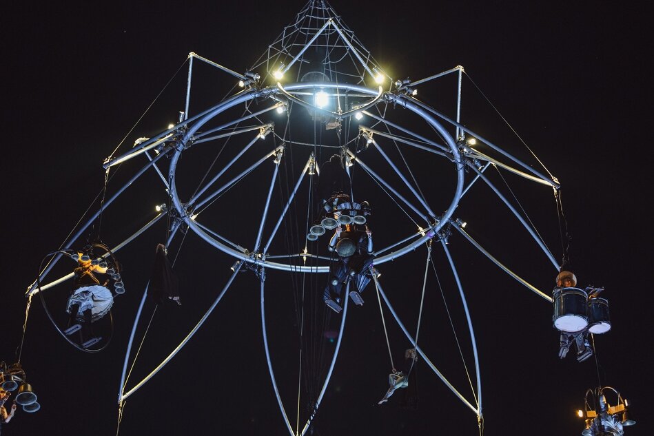 Zdjęcie przedstawia imponującą konstrukcję scenograficzną, zawieszoną wysoko nad ziemią, na której kilku artystów wykonuje akrobacje oraz gra na instrumentach. Konstrukcja jest podświetlona, tworząc spektakularny efekt wizualny na tle nocnego nieba