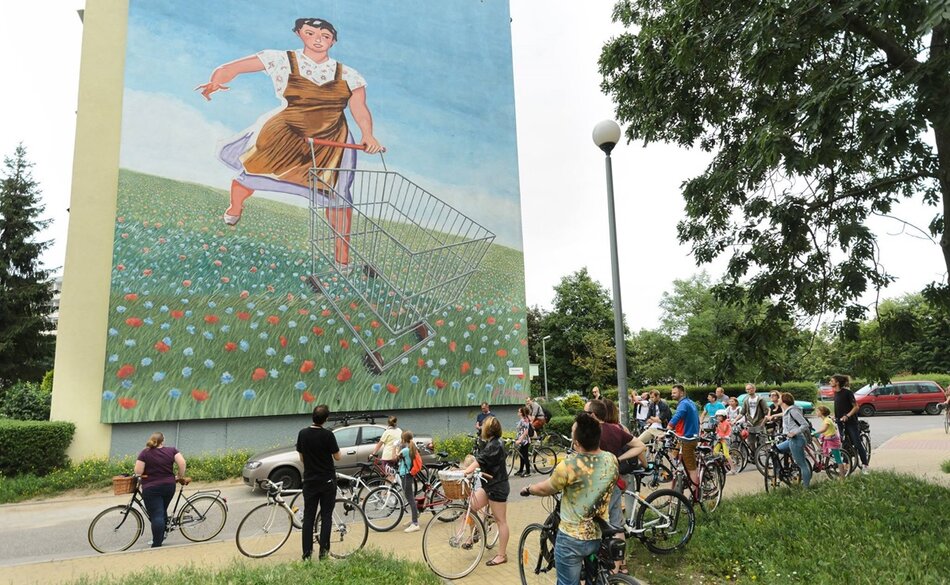 Zdjęcie przedstawia grupę ludzi na rowerach, zatrzymujących się, aby obejrzeć ogromny mural na ścianie budynku. Mural przedstawia kobietę pchającą wózek sklepowy przez pole pełne kwiatów, tworząc kolorowy i dynamiczny widok w miejskiej przestrzeni.