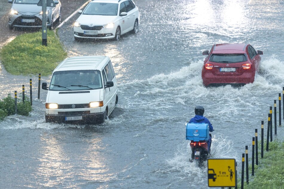 Zdjęcie przedstawia ulicę w trakcie ulewy, która spowodowała znaczne zalanie jezdni. Samochody osobowe, dostawczy Volkswagen oraz skuter z dostawcą jedzenia przemieszczają się przez głęboką wodę, tworząc duże fale.