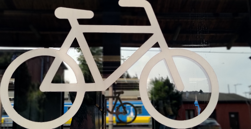 Zdjęcie przedstawia białą ikonę roweru na szklanej powierzchni, za którą widać stację kolejową oraz stojący pociąg w niebiesko-żółtych barwach. W tle znajdują się budynki oraz fragment peronu, co sugeruje, że zdjęcie zostało wykonane na stacji kolejowej.
