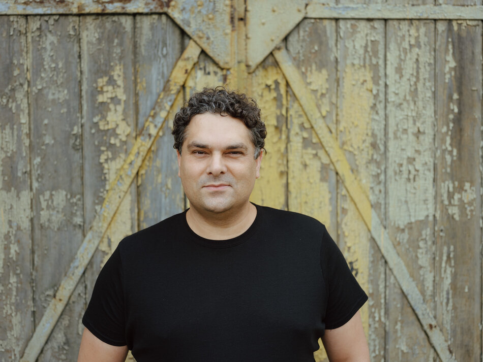 Na zdjęciu widzimy mężczyznę o krótkich, kręconych włosach, ubranego w czarną koszulkę, stojącego na tle starej, drewnianej bramy z odpadającą farbą. Mężczyzna patrzy prosto w obiektyw, a jego wyraz twarzy jest spokojny i pewny siebie.
