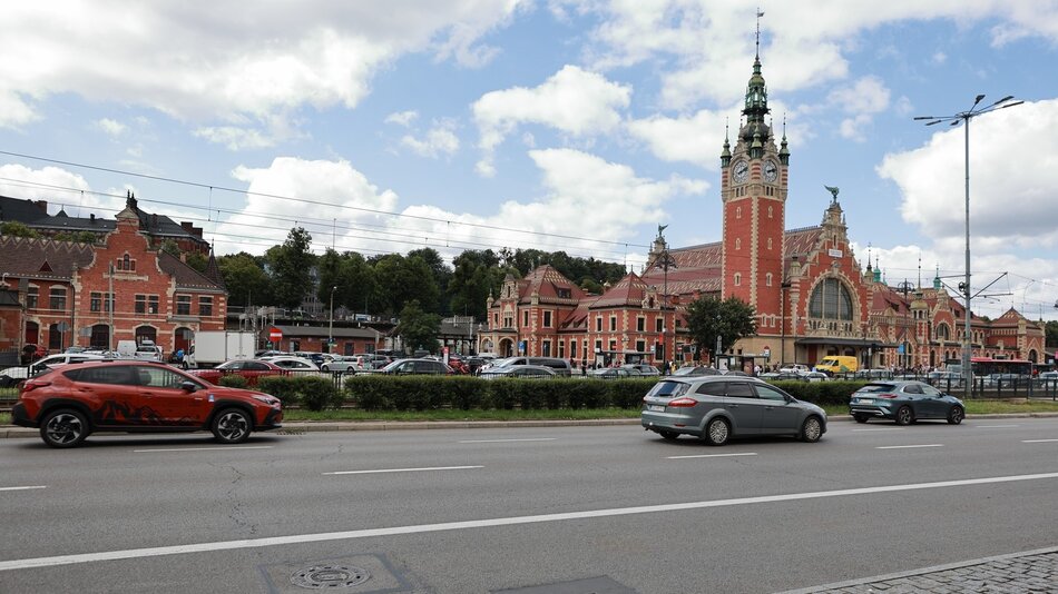 Zdjęcie przedstawia widok na charakterystyczną, zabytkową stację kolejową w dużym mieście, prawdopodobnie europejskim. Budynek stacji wyróżnia się czerwono-ceglaną fasadą, licznymi wieżyczkami oraz dużym zegarem na jednej z wież. Przed stacją znajduje się ruchliwa ulica z kilkoma samochodami w ruchu. Po lewej stronie zdjęcia widać budynki w podobnym stylu architektonicznym, również wykonane z czerwonej cegły. W tle, za stacją, znajduje się zieleń, sugerująca obecność parku lub lasu. Pogoda jest słoneczna z lekkim zachmurzeniem, co dodaje zdjęciu jasnych i ciepłych tonów.