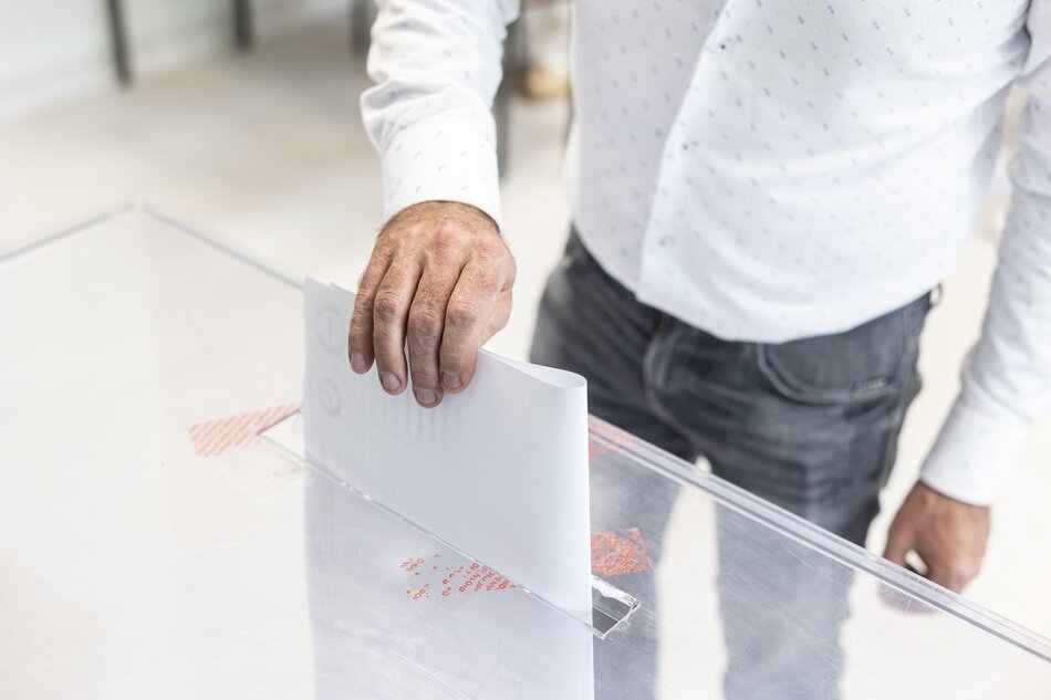 na zdjęciu przezroczysta urna wyborcza, ludzka dłoń wsuwa kartę do głosowania do tej urny