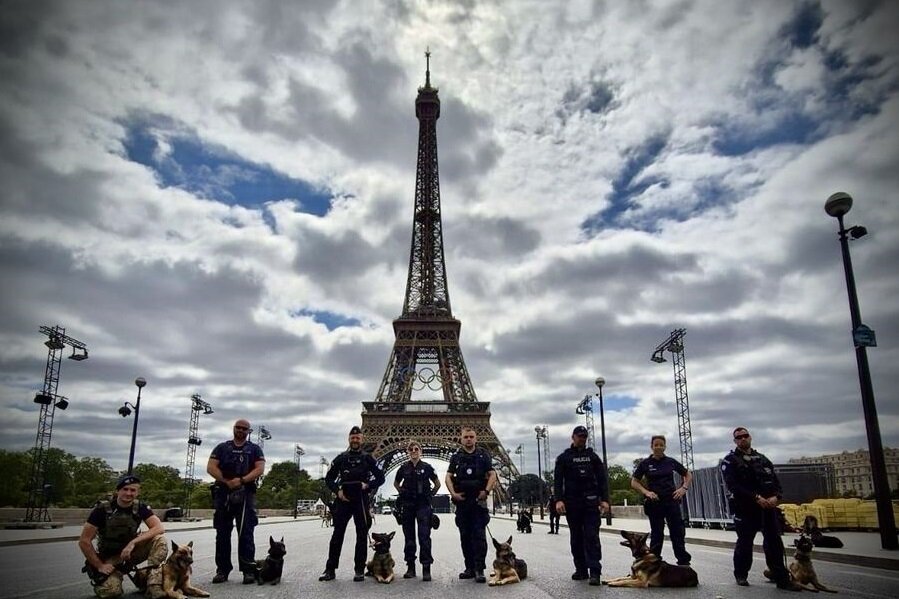 Zdjęcie przedstawia grupę funkcjonariuszy policji wraz z psami policyjnymi, stojących na tle Wieży Eiffla w Paryżu. Siedmiu policjantów, niektórzy w pozycji stojącej, inni klęczący, pozują do zdjęcia na pustej, szerokiej ulicy, co nadaje obrazowi majestatyczny charakter.