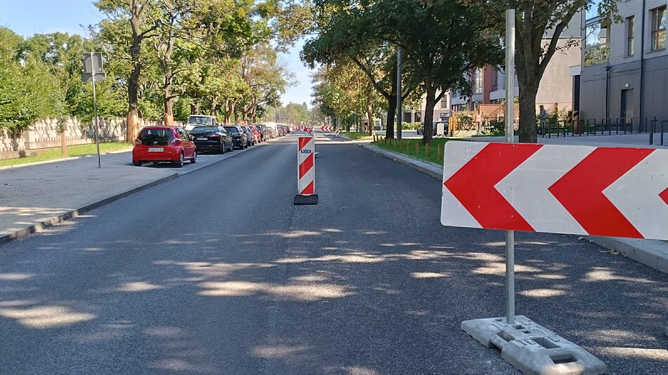 Na zdjęciu widoczna jest ulica, która jest częściowo zamknięta z powodu prac drogowych. Wzdłuż drogi ustawione są biało-czerwone bariery i pachołki, które kierują ruch na jeden pas. W tle można zauważyć kolejne bariery, co wskazuje na to, że prace drogowe obejmują dłuższy odcinek drogi. Po lewej stronie ulicy widać zaparkowane samochody, a wzdłuż chodnika rosną drzewa, które zapewniają cień. Po prawej stronie ulicy znajdują się budynki mieszkalne oraz chodnik. Pogoda wydaje się być słoneczna, co można wywnioskować z cieni rzucanych przez drzewa. Cała scena wygląda na spokojną, a na ulicy nie widać aktywnego ruchu pojazdów.