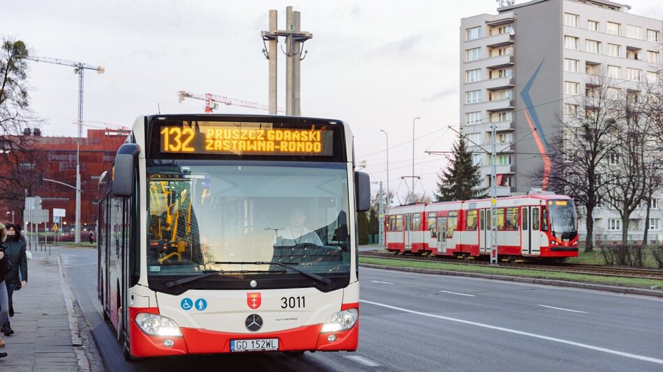 Na zdjęciu widoczny jest autobus miejski linii 132, który jedzie w kierunku Pruszcza Gdańskiego, z przystankiem końcowym Zastawna-Rondo. Autobus jest biało-czerwony, marki Mercedes-Benz, i ma numer rejestracyjny GD 152WL. Z przodu autobusu widoczne są piktogramy informujące o dostępności dla osób niepełnosprawnych oraz dla wózków dziecięcych. Po prawej stronie zdjęcia widać również tramwaj w barwach biało-czerwonych, jadący po torowisku równoległym do ulicy. W tle znajdują się wysokie budynki mieszkalne, z jednym z nich posiadającym charakterystyczny mural w kształcie strzały. Na dalszym planie widoczne są żurawie budowlane, co sugeruje, że w okolicy prowadzone są prace budowlane.