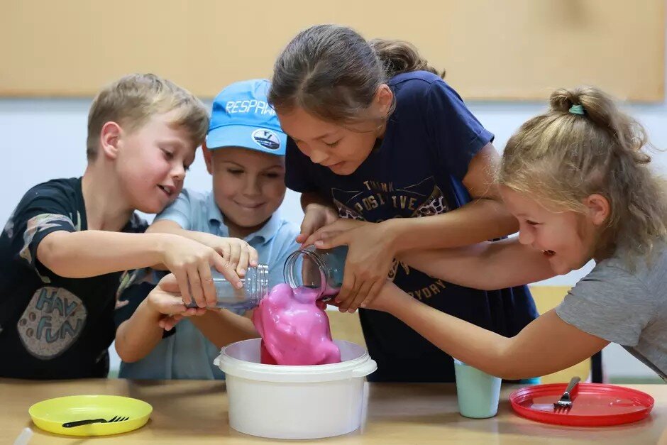 Na zdjęciu widać czworo dzieci zaangażowanych w eksperyment chemiczny, polegający na tworzeniu piany w misce. Dzieci wydają się być zadowolone i uśmiechnięte, współpracując ze sobą podczas zabawy.