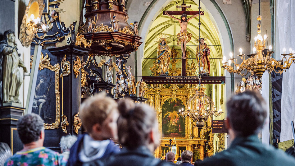 Wnętrze zabytkowego kościoła wypełnione bogato zdobioną stolarką. Na pierwszym planie grupa ludzi, w tle widoczny pozłacany ołtarz