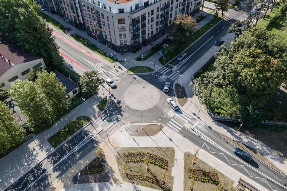 Na zdjęciu przedstawiono rondo w centrum miasta, otoczone nowoczesnymi budynkami mieszkalnymi oraz zielenią miejską. Rondo posiada oznakowane przejścia dla pieszych, ścieżki rowerowe oraz jest w pełni zintegrowane z otaczającą infrastrukturą drogową.