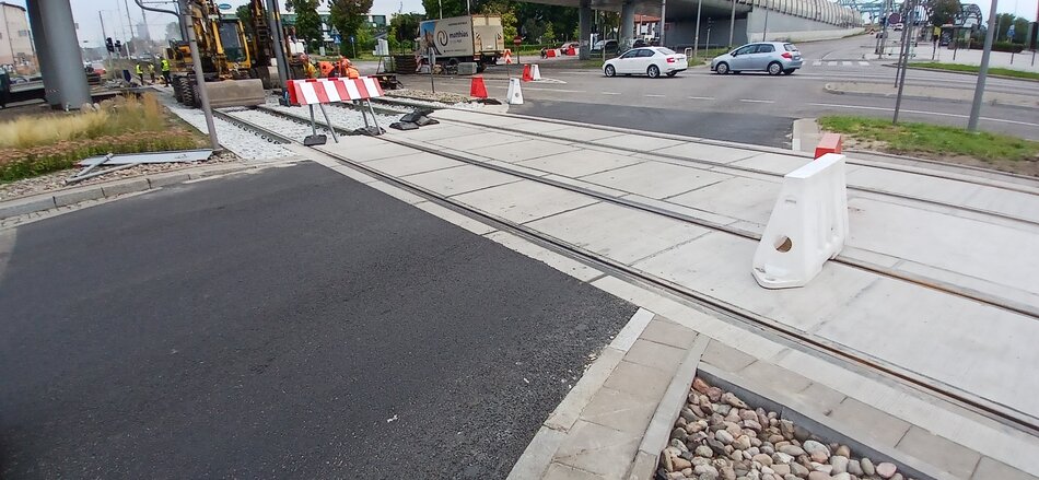 Na zdjęciu widać remontowany przejazd tramwajowy, gdzie pracownicy zajmują się układaniem nowych torów oraz nawierzchni. Obszar jest częściowo odgrodzony barierkami i pachołkami, a w tle widoczne są maszyny budowlane oraz samochody na drodze.