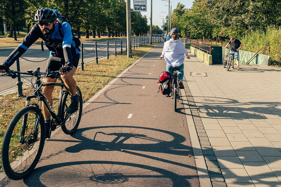 Po ścieżce rowerowej w mieście jedzie mężczyzna w stroju sportowym i kobieta w stroju codziennym z sakwą. Na chodniku stoi mężczyzna z rowerem. Wokół rosną drzewa.