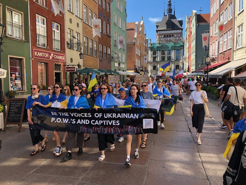 Na zdjęciu widać marsz solidarności z Ukrainą, który odbywa się na ulicy Długiej w Gdańsku. Uczestnicy marszu niosą duży czarny baner z napisem w języku angielskim: BE THE VOICES OF UKRAINIAN P.O.W.S AND CAPTIVES, co oznacza Bądźcie głosem ukraińskich jeńców wojennych i więźniów. Wiele osób ma na sobie niebiesko-żółte flagi lub chusty, co symbolizuje wsparcie dla Ukrainy. W tle widzimy kolorowe, zabytkowe kamienice Gdańska oraz Złotą Bramę, charakterystyczny punkt architektoniczny miasta. Na transparentach widać także napisy w języku polskim i angielskim, dziękujące Polsce za wsparcie dla Ukrainy. Atmosfera jest spokojna i pełna solidarności, a na ulicach widać wiele ludzi obserwujących marsz lub biorących w nim udział. Pogoda jest słoneczna, co widać po jasnym oświetleniu i cieniu rzucanym przez uczestników marszu.