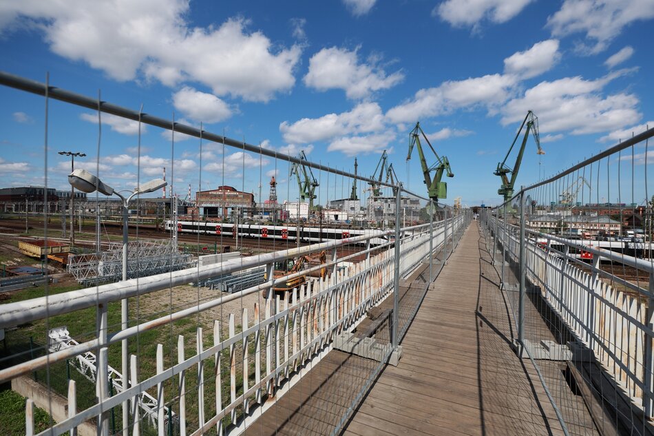Drewniana, zniszczona kładka dla pieszych nad torami kolejowymi. W tle dźwigi portowe.