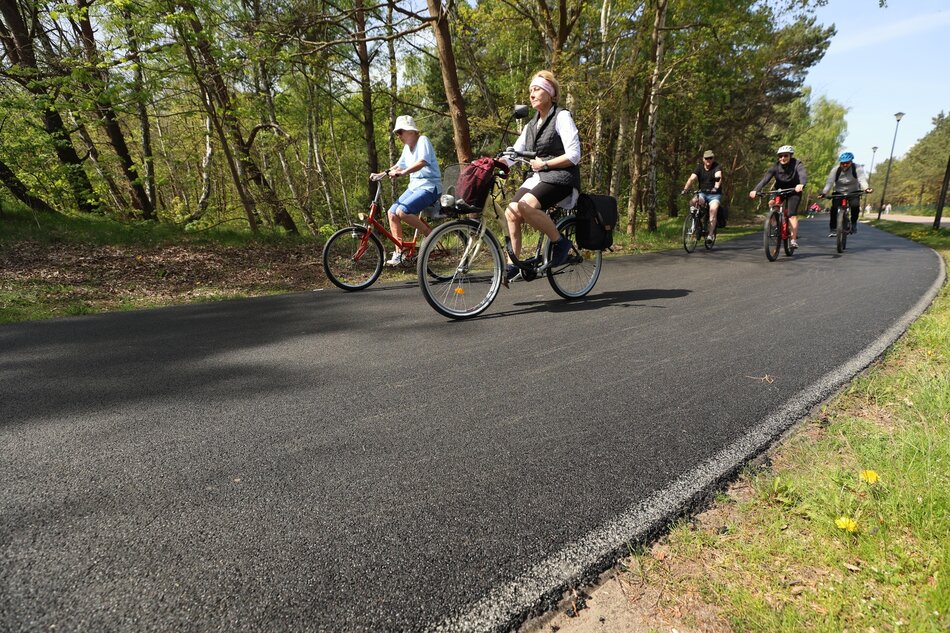 Zdjęcie przedstawia grupę rowerzystów jadących nowo wyasfaltowaną ścieżką rowerową w lesie, otoczoną zielonymi drzewami. Pogoda jest słoneczna, a rowerzyści wydają się cieszyć jazdą, co sugeruje relaksacyjną, rekreacyjną przejażdżkę w naturalnym otoczeniu.