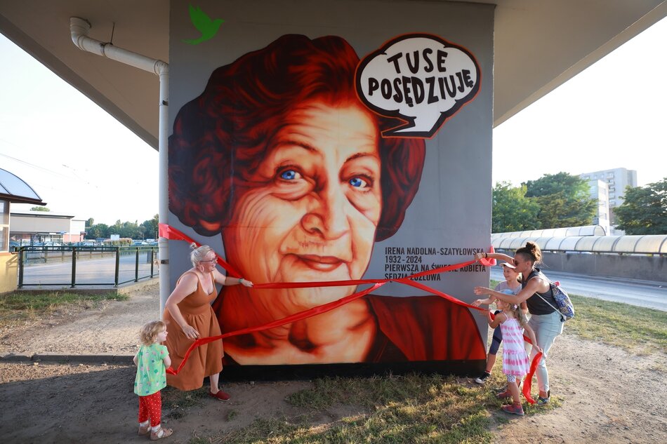 na zdjęciu mural przedstawiający twarz starszej kobiety, w brązowo czerwonych odcieniach, mural odsłaniają dwie kobiety i dzieci, które ściągają z muralu dużą czerwoną wstążkę