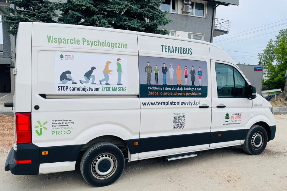 Zdjęcie przedstawia biały samochód oznaczony jako Terapiobus, który oferuje wsparcie psychologiczne. Na bocznej ścianie pojazdu znajduje się plakat promujący zapobieganie samobójstwom oraz hasła zachęcające do dbania o zdrowie psychiczne, z informacją o stronie internetowej kampanii terapiatoniewstyd.pl.