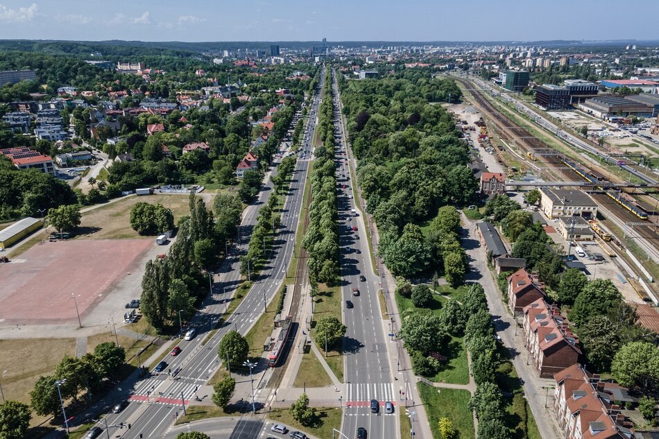 Zdjęcie przedstawia widok z lotu ptaka na zieloną okolicę miejską z szeroką ulicą pośrodku, którą oddziela pas drzew, a obok biegnie linia kolejowa z peronami i torami. Na dalszym planie widoczne są zarówno budynki mieszkalne, jak i przemysłowe, a także rozciągający się horyzont miasta.