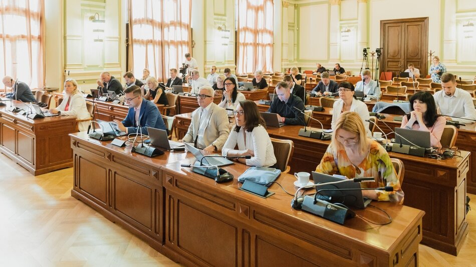 Na zdjęciu widać formalne spotkanie lub konferencję odbywającą się w eleganckiej, dobrze oświetlonej sali z wysokimi oknami. Pomieszczenie jest urządzone w klasycznym stylu, z wysokimi sufitami, zdobionymi sztukateriami i dużymi, drewnianymi biurkami ustawionymi w rzędach. Uczestnicy, zarówno mężczyźni, jak i kobiety, siedzą przy tych biurkach, z których każde jest wyposażone w laptopy, mikrofony oraz dokumenty. To wskazuje na oficjalny charakter wydarzenia, prawdopodobnie jest to posiedzenie rady, konferencja lub warsztaty. Uczestnicy wydają się zaangażowani, niektórzy robią notatki, inni uważnie słuchają lub korzystają z laptopów. Cała atmosfera sugeruje skupienie na dyskusji, planowaniu lub podejmowaniu decyzji.